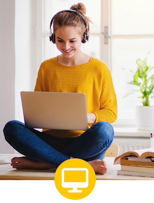 Junge Frau im gelben Pullover und Jeans sitzt im Schneidersitz auf einem Tisch neben einem kleinen Bücherstapel, auf ihrem Schoß hat sie einen Laptop und sie trägt Kopfhörer