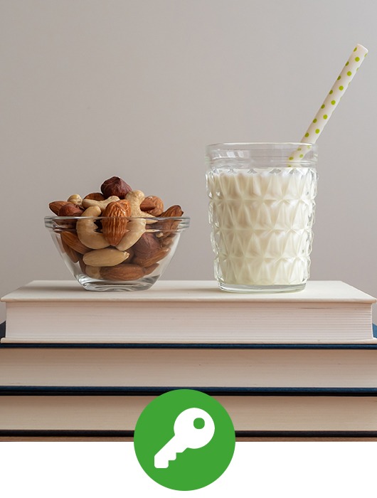 Links eine kleine Glasschüssel gefüllt mit Studentenfutter und rechts ein Glas Milch mit Strohhalm auf einem Bücherstapel mit drei Büchern